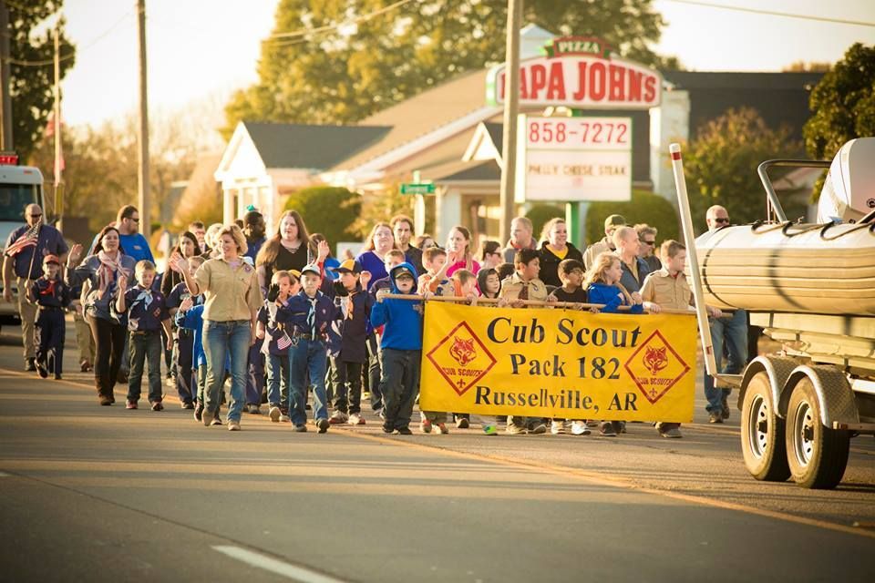 Scout Pack 182 parade marchers carry a yellow banner beside a trailer on a sunny street.