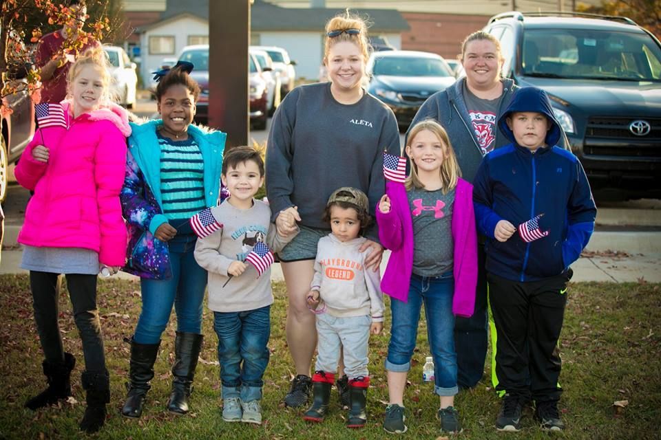 Seven children standing together outdoors in front of parked vehicles, wearing colorful jackets in daylight.