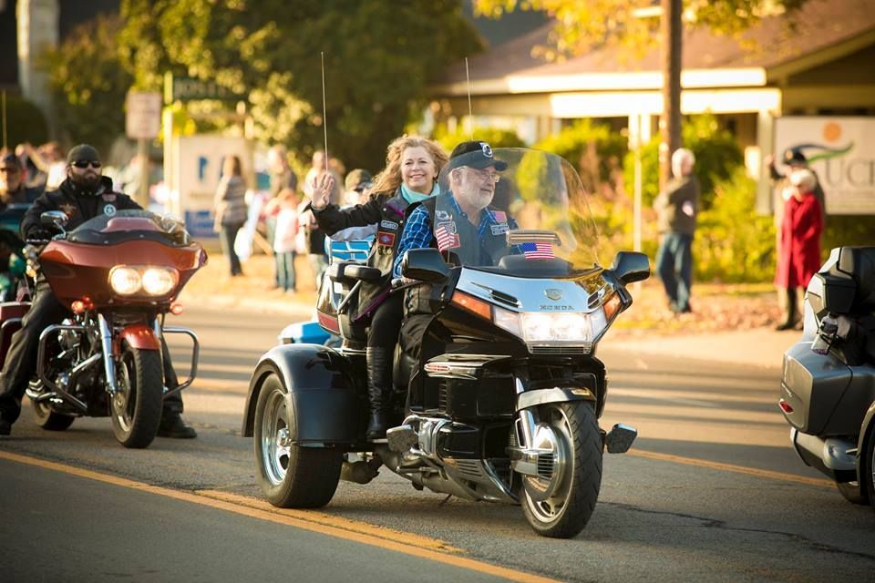 Trike riders and motorcyclists cruise down a sunny street, one rider waving to the crowd.