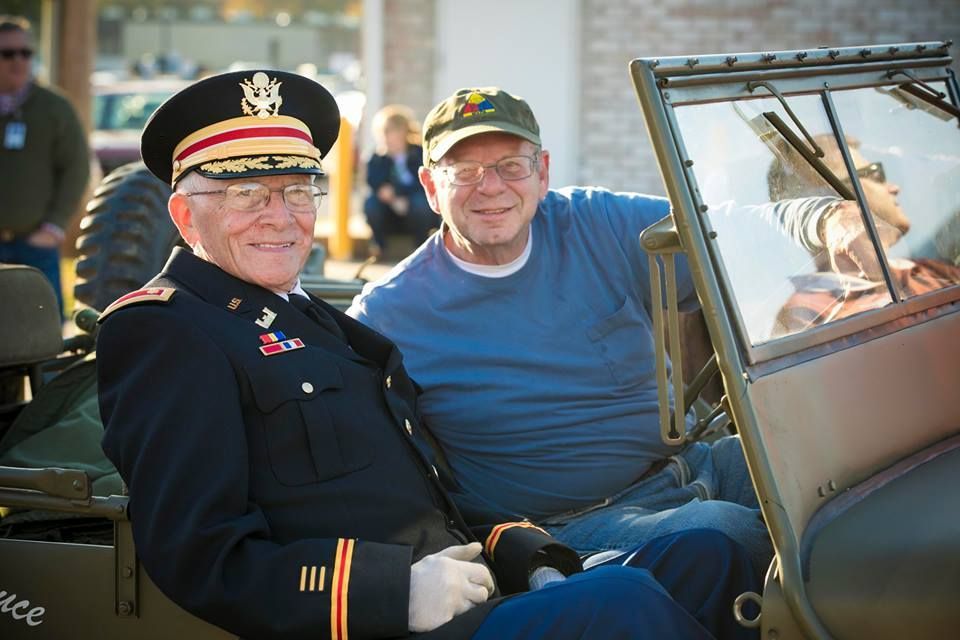 Two smiling men seated in a vintage open car, one in a dark military uniform and cap, outdoors.