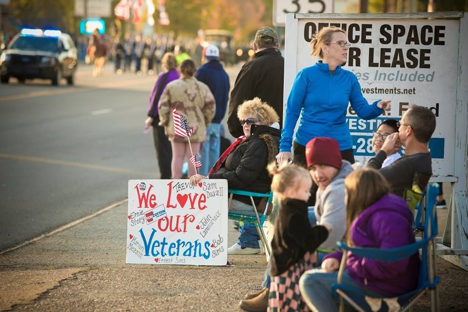 People sitting along a roadside holding a sign that reads, “We Love Our Veterans”