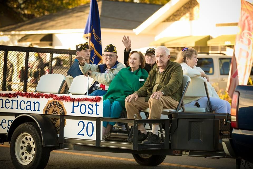 Veterans riding in a parade float with an American Legion Post 20 sign and flags at sunset