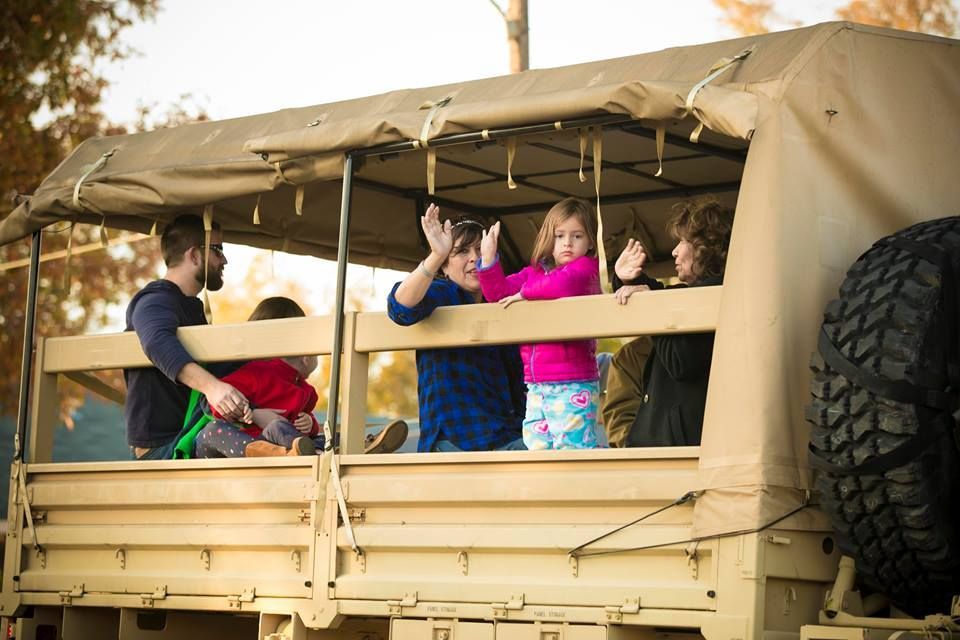 People waving from the back of a tan safari-style truck with a spare tire on the side.