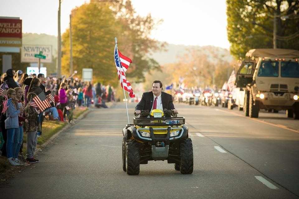 Man riding an ATV in a parade with a flag, crowds lining the street and a military vehicle behind him