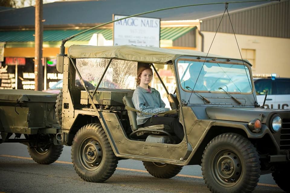 Open-top military jeep with a driver parked in a market area, towing a small trailer.
