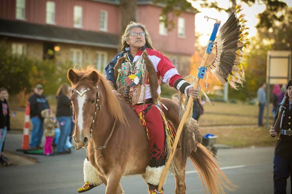 Person in colorful regalia riding a pony in a parade, holding a feather fan on a street with spectators