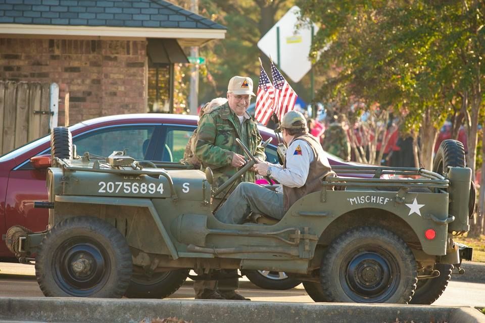 Veteran in camouflage uniform riding in a green military jeep, waving with an American flag in a street parade