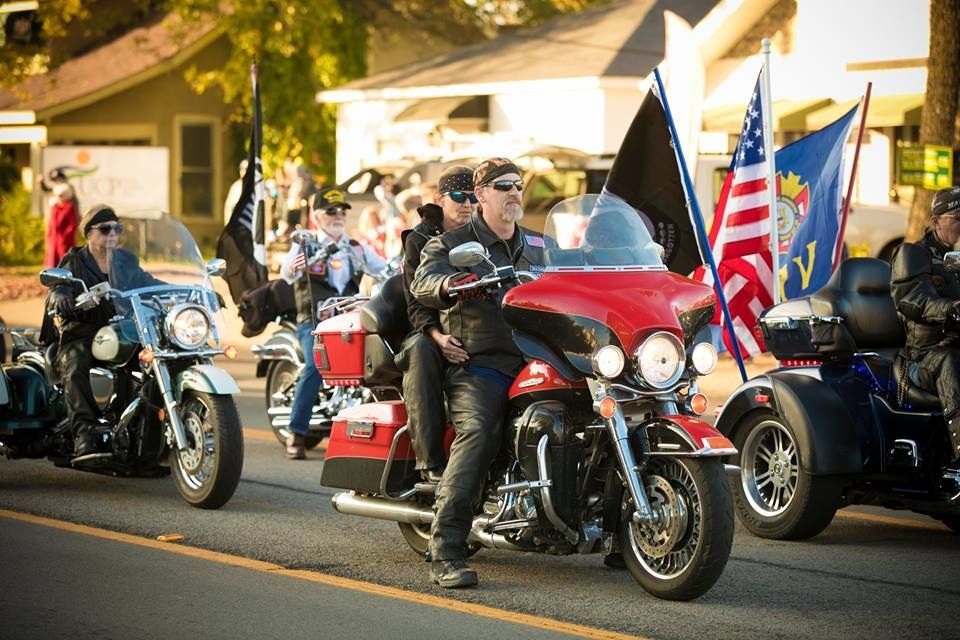 Motorcyclists in a parade ride down a sunlit street, with American flags and colorful banners.