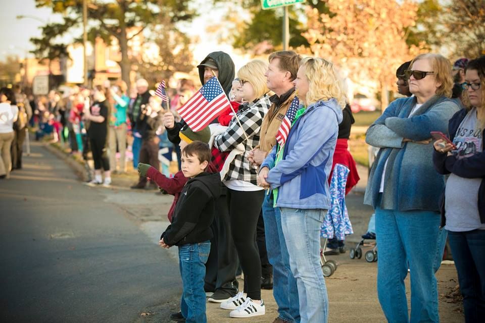 People standing along a sidewalk at a street event, some holding American flags, in autumn daylight