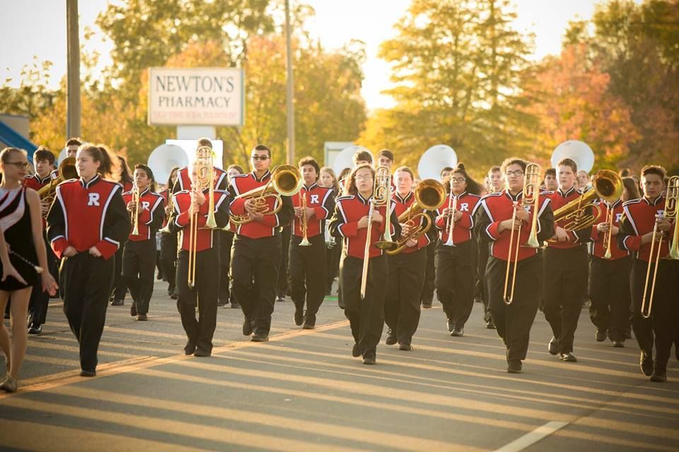 Marching band in red and black uniforms performs on a sunlit street during a parade