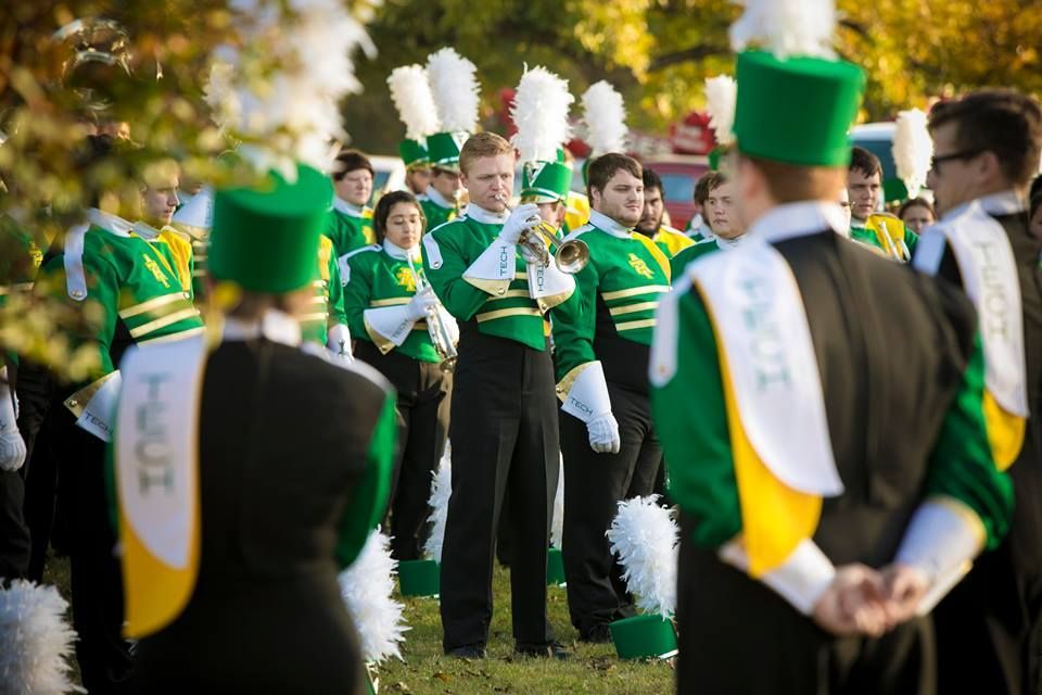 Band members in green and white uniforms standing outdoors, some wearing tall green hats.