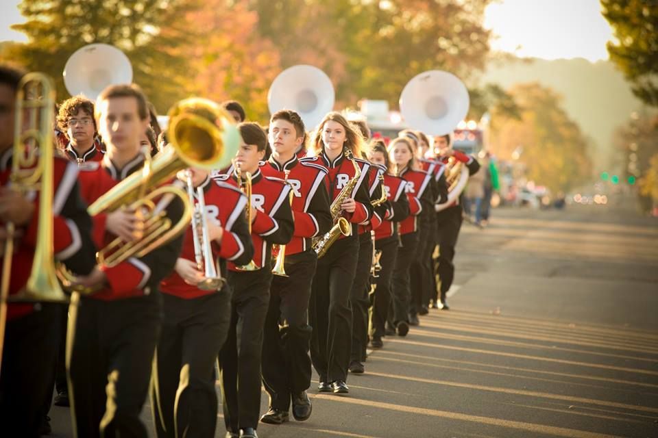 Marching band in red uniforms playing brass instruments in a parade line on a sunlit street.