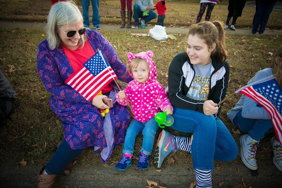 Woman and two children seated outdoors, holding small American flags at a patriotic event.