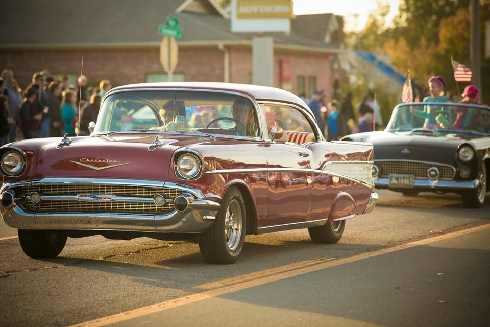 Classic maroon and white vintage car driving in a sunny parade with other classic cars behind it