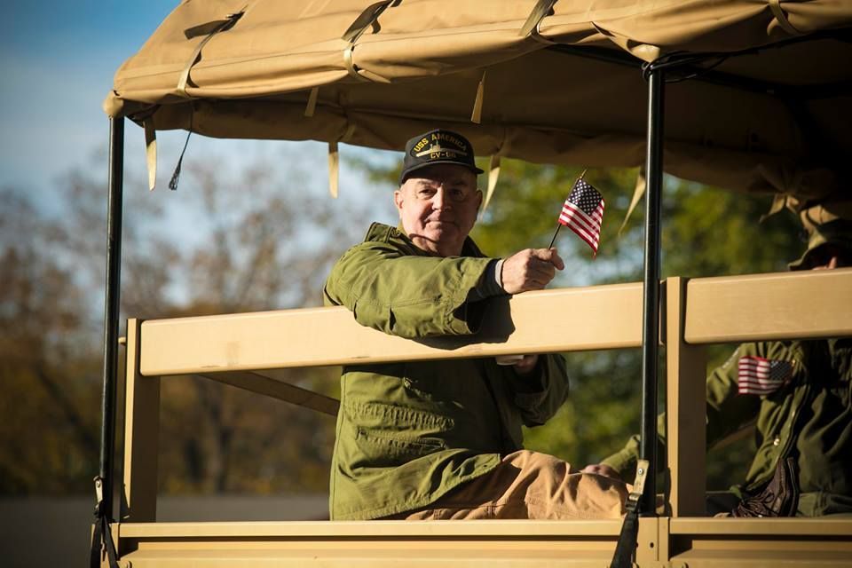 Person in green military uniform waving a small American flag from an open-sided vehicle.