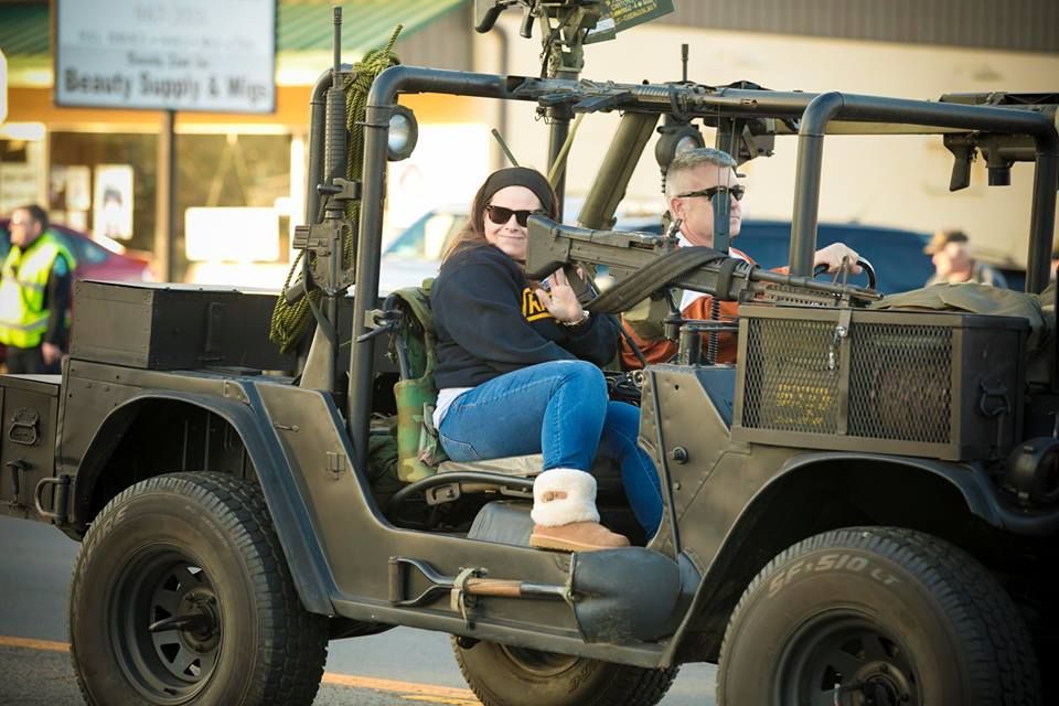 Two people ride in an open-top military-style jeep, one driving and one seated beside him, outdoors.