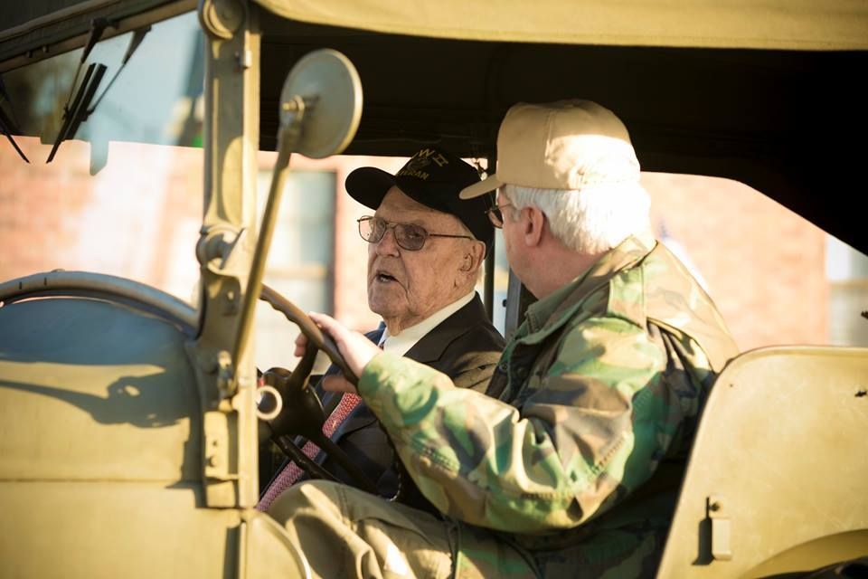 Two men in an open-top vintage vehicle, one driving, both wearing caps under a canopy.