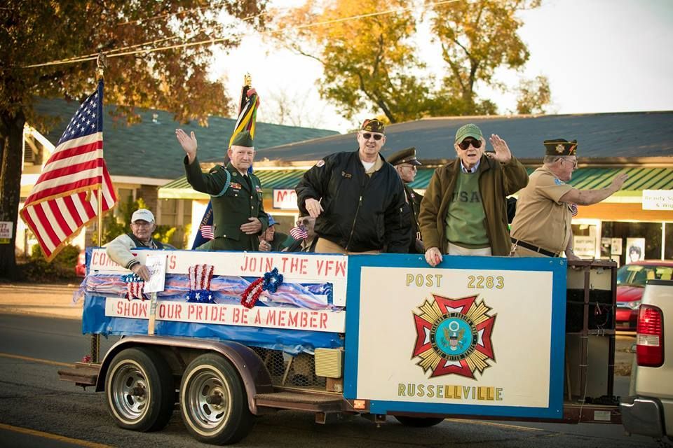 Veterans on a parade float with American flags and a Russellville post sign, waving outdoors