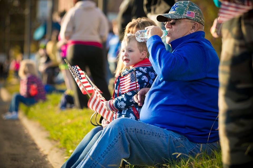 Spectators seated on grass at a parade, a child waving a small American flag beside an older man in blue.