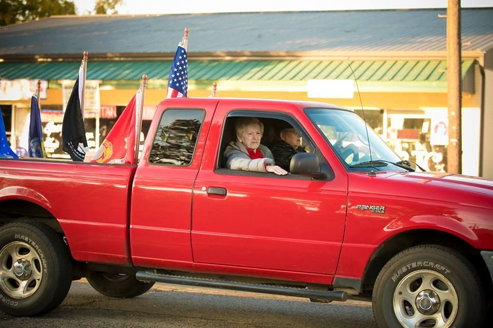 Red pickup truck with two children in the cab, parked outside a building with flags.