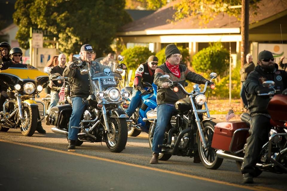 Motorcyclists lined up on a sunlit street, with chrome bikes and riders in helmets near a house-lined road
