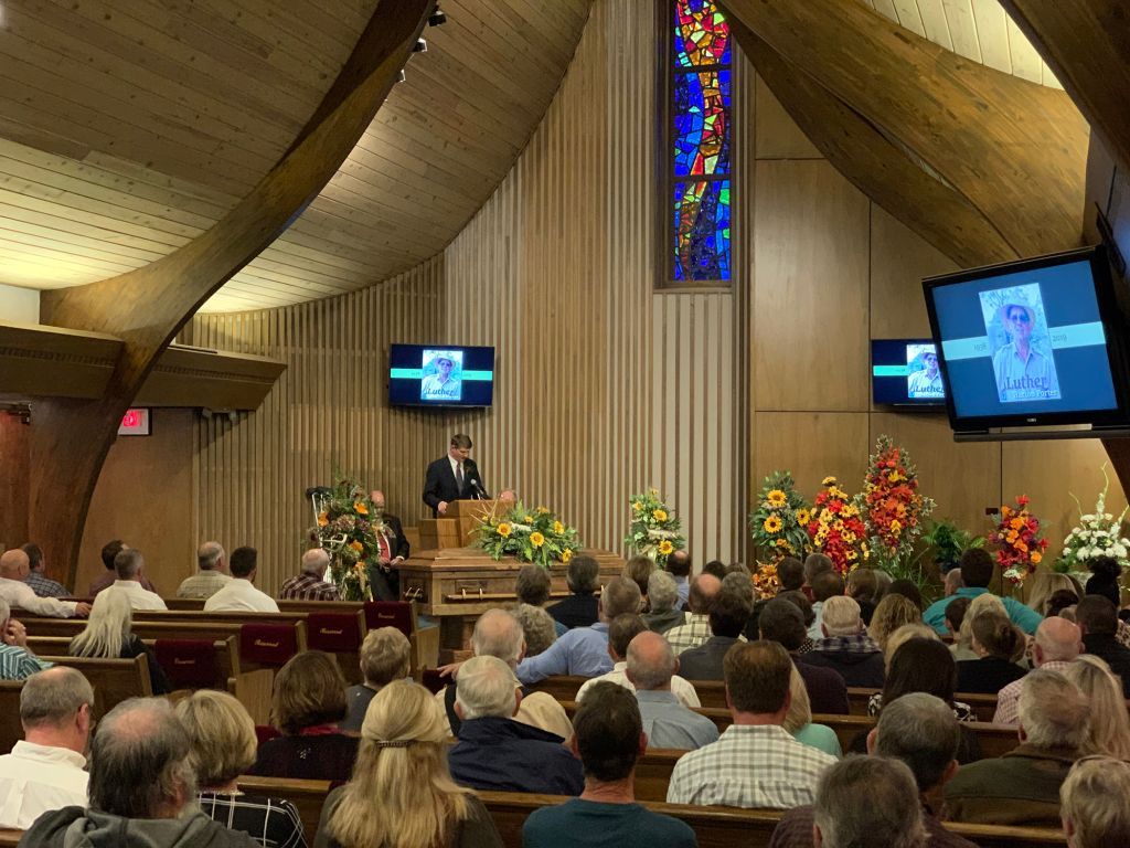 Congregation seated in a church sanctuary during a service, with a speaker at the pulpit and stained-glass window above.