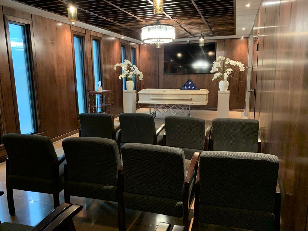 Wood-paneled chapel with rows of black chairs facing a white coffin and floral arrangements