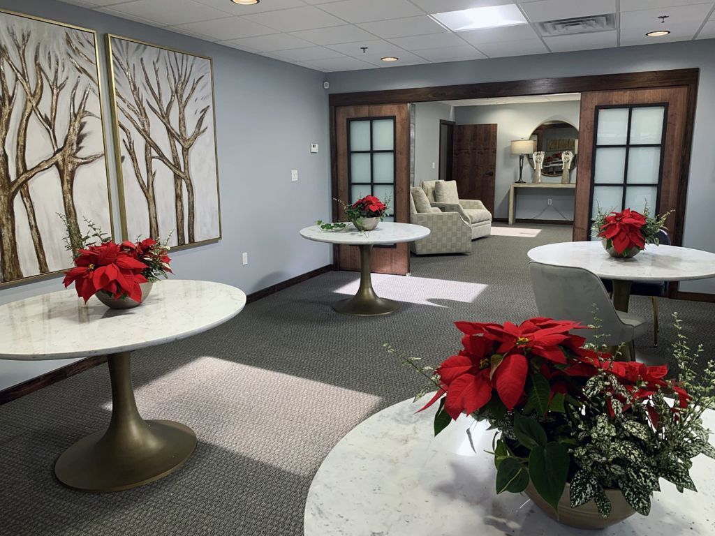 Office lobby with white tables, red poinsettias, and wall art of trees, leading to a seating area.