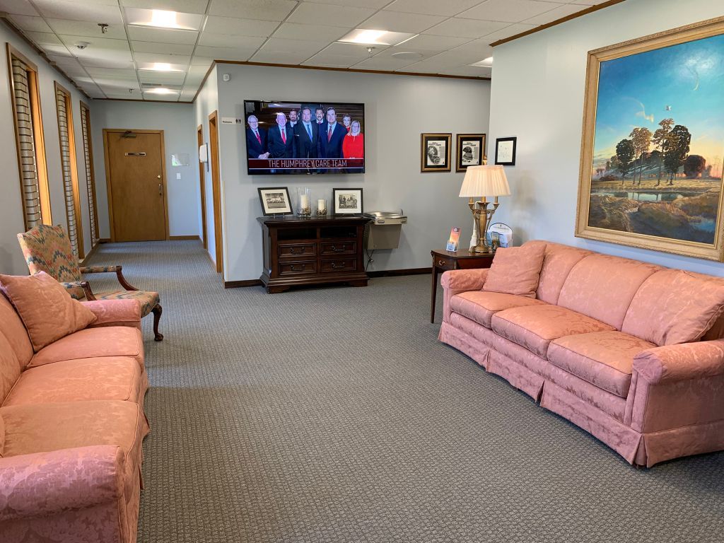 Waiting room with pink sofas, TV, hallway, and framed art in a bright office corridor.