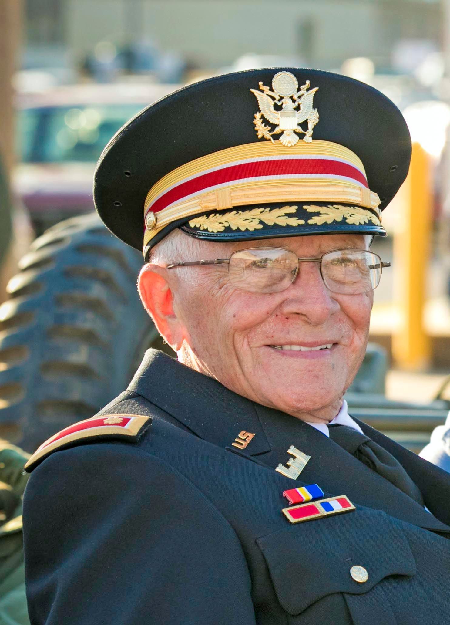Smiling man in a dark military dress uniform and peaked cap, seated outdoors