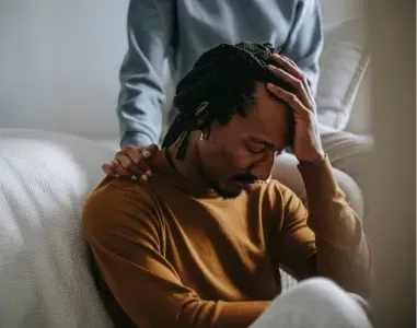 A grieving person sitting with their head in their hand while another person offers comforting support.