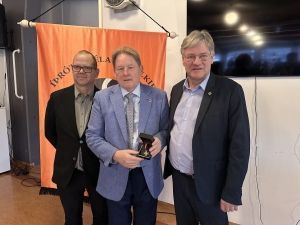 Þrír menn standa innandyra; miðmaðurinn heldur á bikar fyrir framan appelsínugulan borða.
