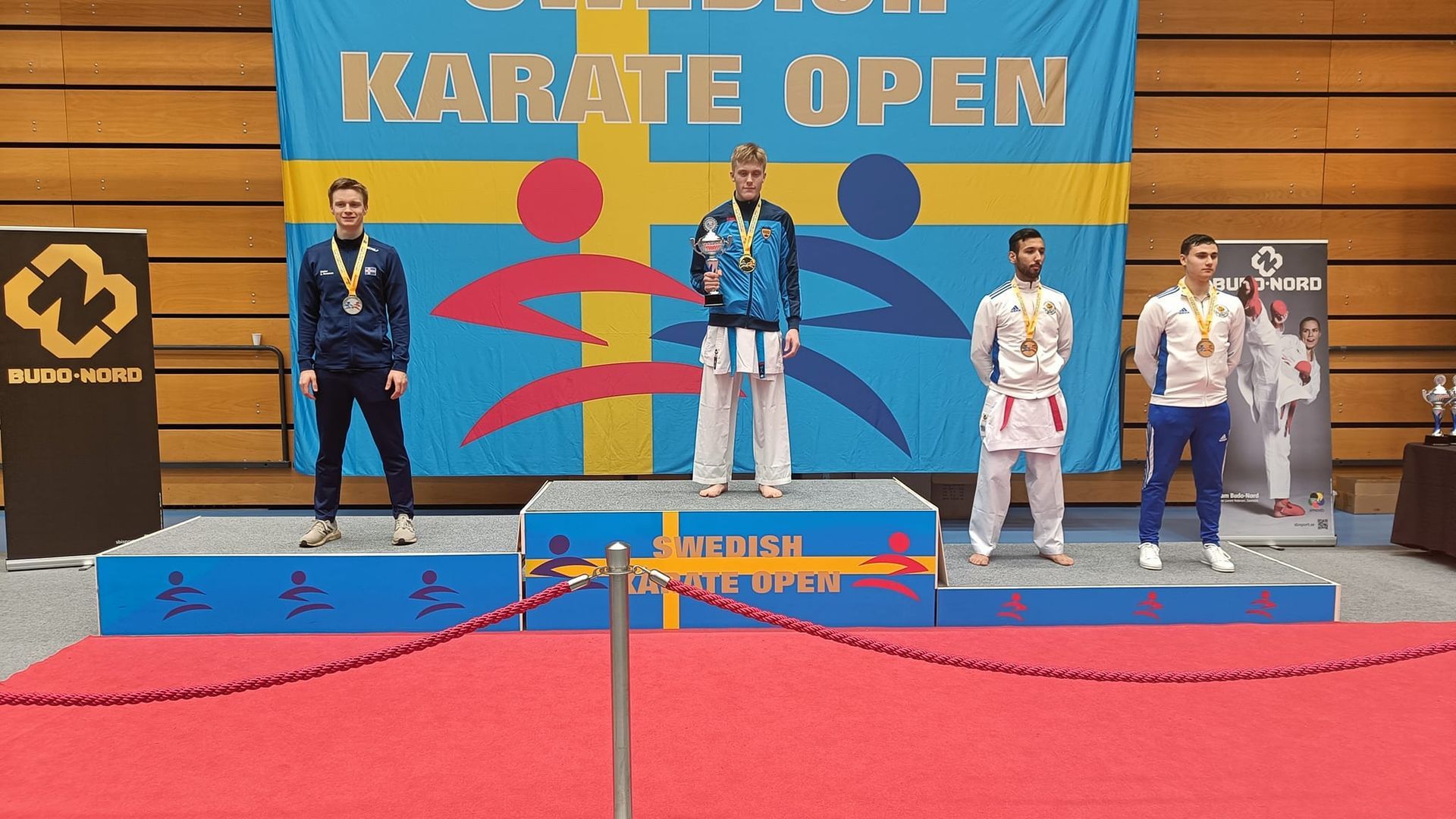Þrír sigurvegarar í bardagaíþróttum á verðlaunapalli í Karate Open móti, með verðlaunapeninga og fána á bak við sig.