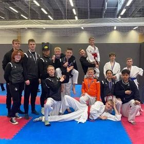 Hópur bardagaíþróttanema sitja fyrir í íþróttahúsi á bláum og rauðum dýnum, sumir í einkennisbúningum og með belti.