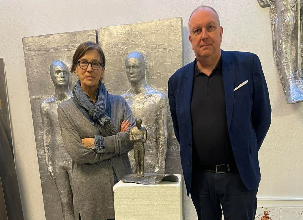 Tveir einstaklingar standa við hlið silfurskúlptúra ​​í galleríi, með krosslagða hendur og horfa í myndavélina.