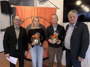 Fjórir einstaklingar sitja fyrir innandyra með blómvönd fyrir framan appelsínugulan borða og vegghengt sjónvarp.