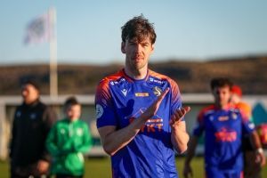 Knattspyrnumaður í bláum treyju klappar á vellinum með liðsfélaga óskýran í bakgrunni.