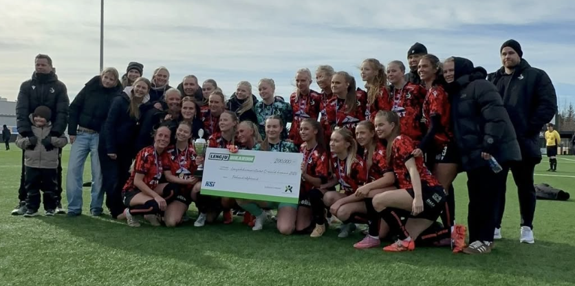 Unglingaíþróttalið poserar á vellinum með stóran verðlaunapening, umkringt þjálfurum og stuðningsmönnum.