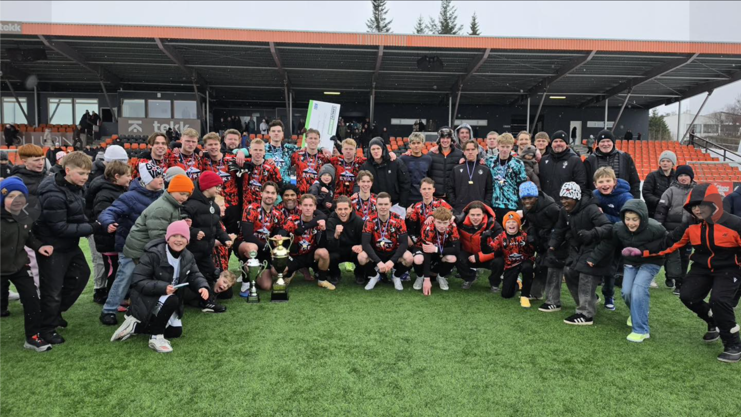 Stór hópur stillir sér upp á knattspyrnuvelli, margir í rauðum og svörtum búningum, með áhorfendur í stúkunni fyrir aftan sig.