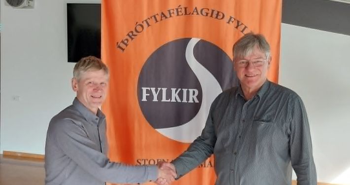 Tveir menn taka í hendur fyrir framan appelsínugulan og svartan fána FYKIR félagsins.