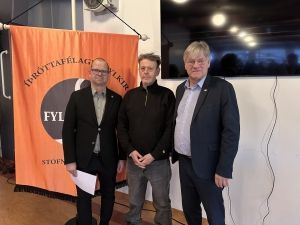 Þrír menn standa innandyra við hliðina á appelsínugulum borða og sjónvarpi sem fest er á vegg.