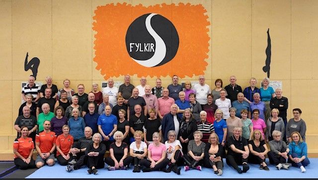 Stór hópur poserar innandyra fyrir framan appelsínugulan vegg með svart-hvítum borða með merki.