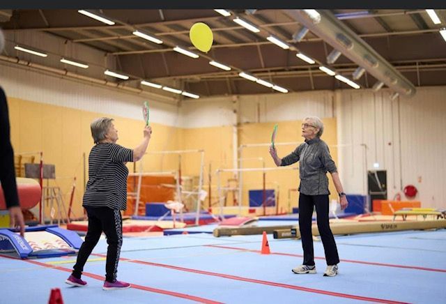 Tveir einstaklingar spila blak í íþróttahúsi og teygja sig eftir gulum bolta á bláum dýnugólfinu.