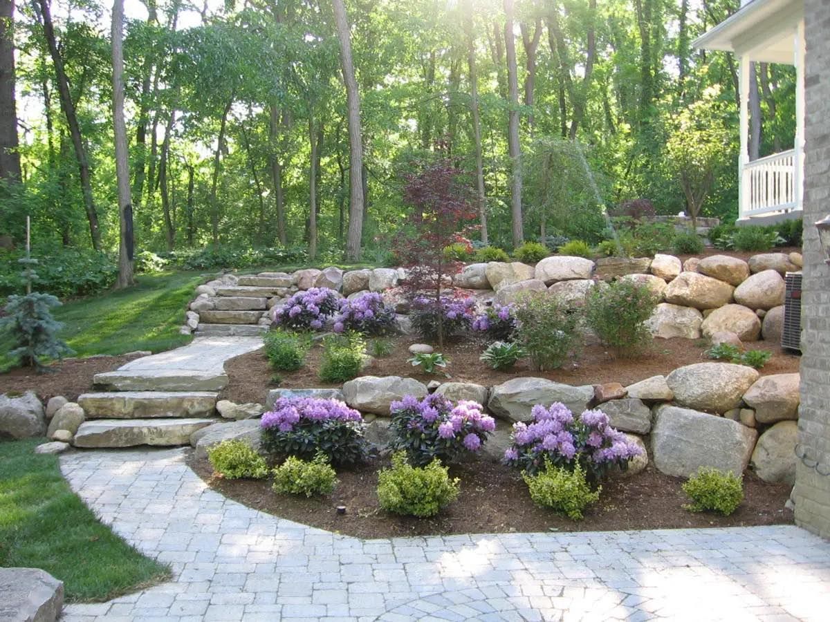 Stone garden path with purple flowers, retaining wall, and wooded backyard patio area