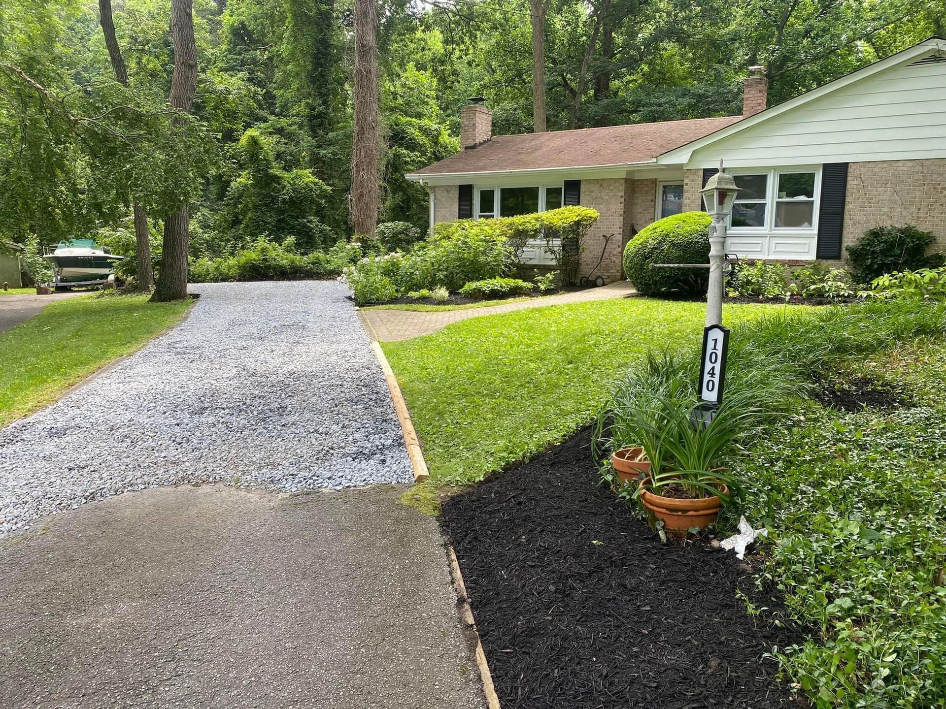 Gravel driveway beside a house with a green lawn, trees, and a porch in a wooded neighborhood