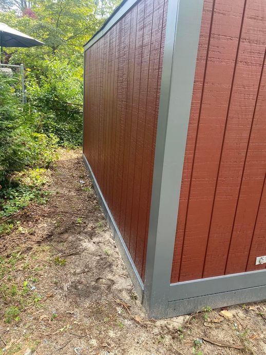 Red-brown wooden shed with gray trim beside a narrow gravel path and green shrubs.