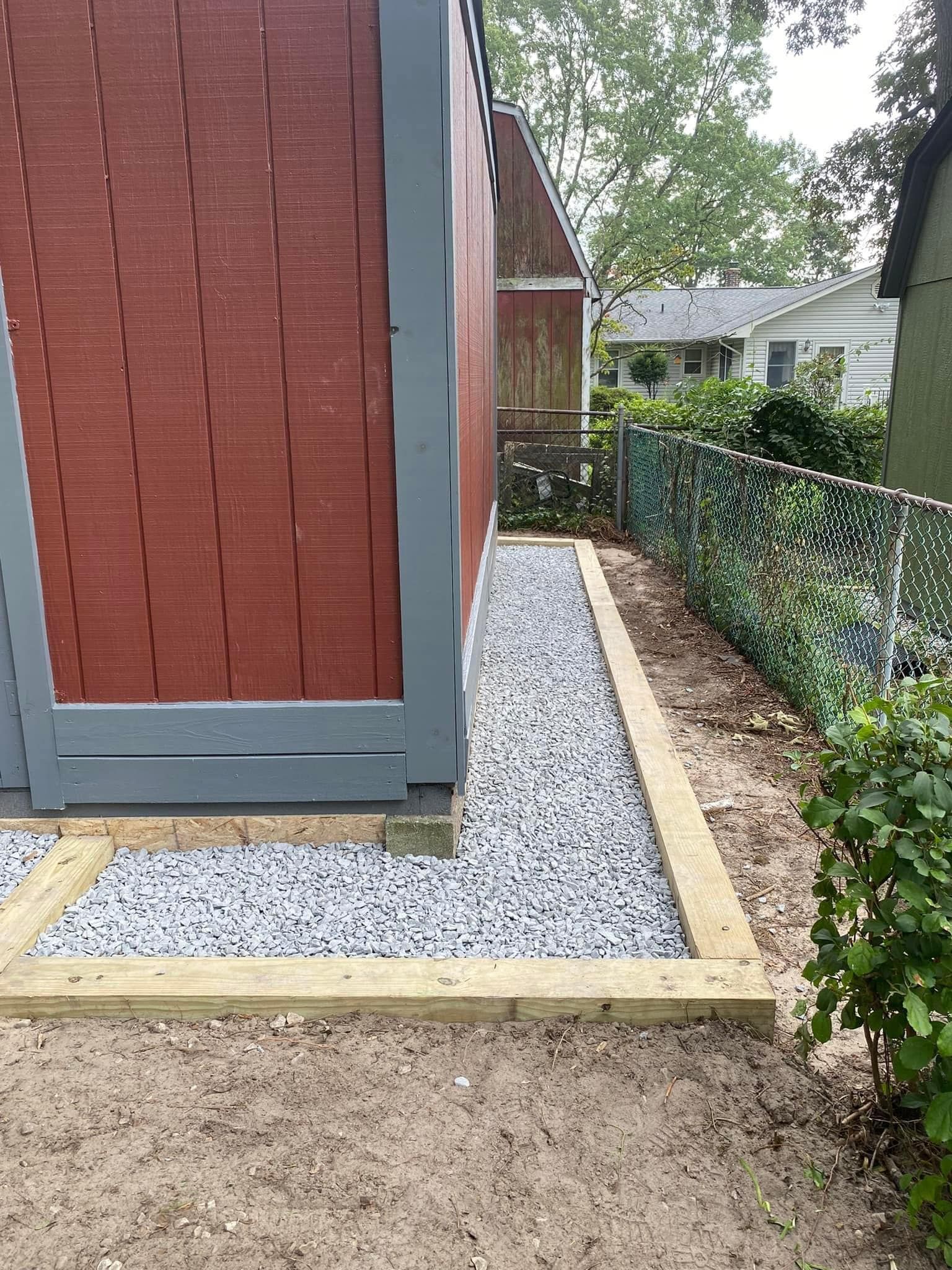 Gravel walkway beside a red shed with gray trim, bordered by wood edging and a chain-link fence.