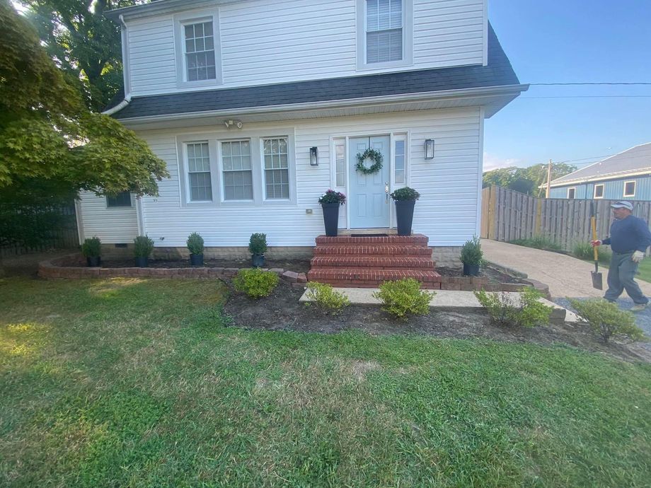 White two-story house with front steps, small garden, and a person walking on the driveway.