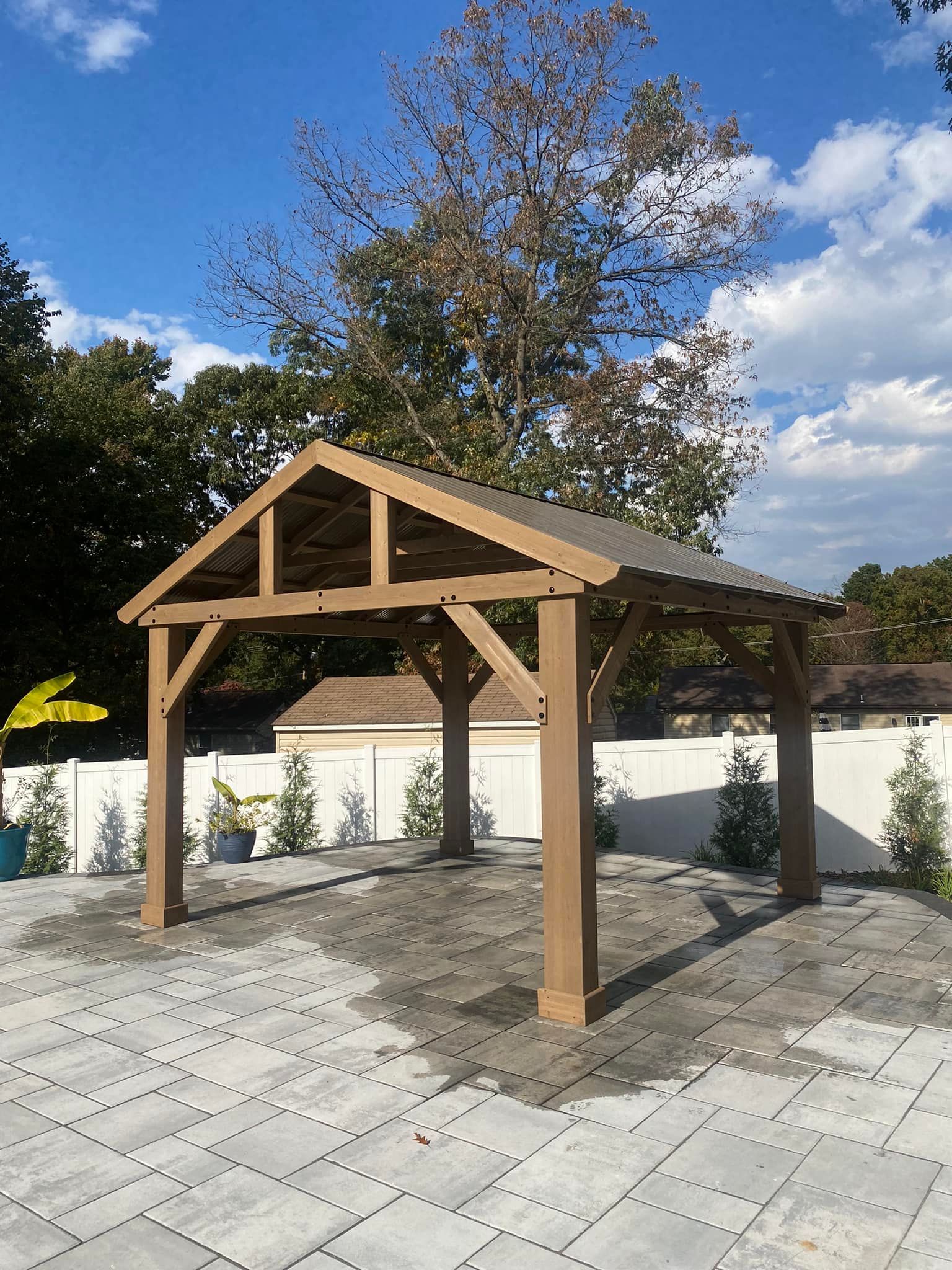 Wooden gazebo on a paved patio, with trees and blue sky in the background.