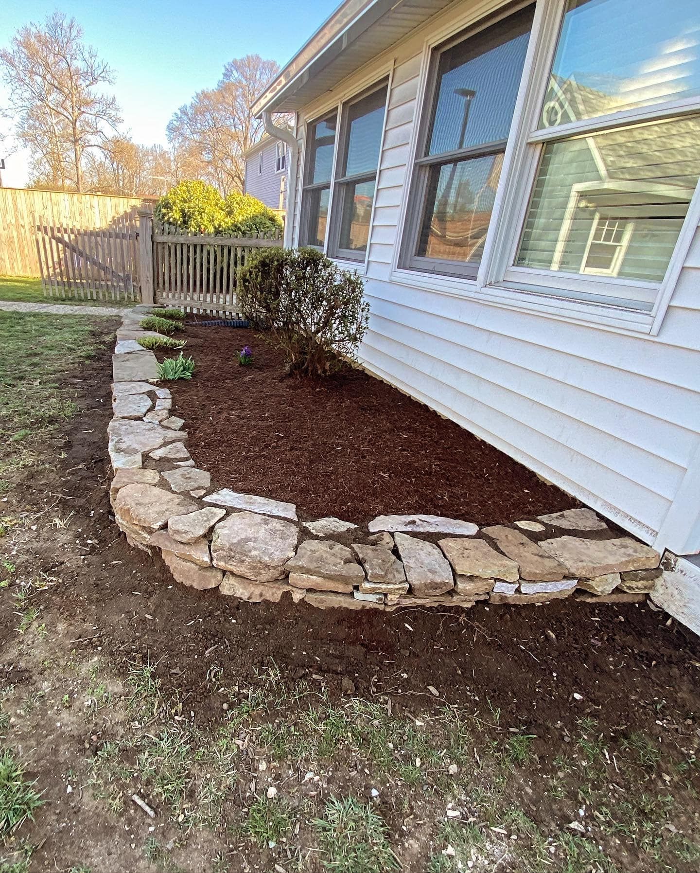 Curved stone border around a mulched garden bed beside a white house with windows